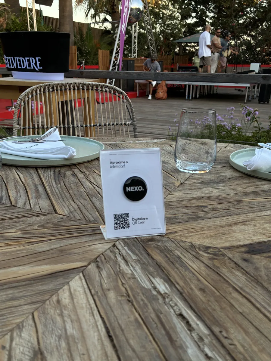 Display NEXO numa mesa de madeira de uma esplanada real: «Aproxime o telemóvel» com o chip NFC e o QR code por baixo.